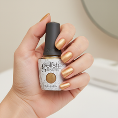 Mano sosteniendo el frasco de esmalte en gel Gelish Give Me Gold, color Nudes acabado Glitter, con las uñas pintadas del mismo tono