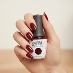 Mano sosteniendo el frasco de esmalte en gel Gelish Red Alert, color Borgoñas acabado Cream, con las uñas pintadas del mismo tono