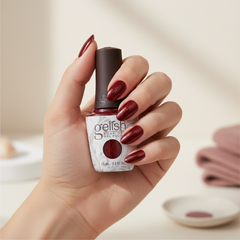Mano sosteniendo el frasco de esmalte en gel Gelish Good Gossip, color Rojos acabado Shimmer, con las uñas pintadas del mismo tono