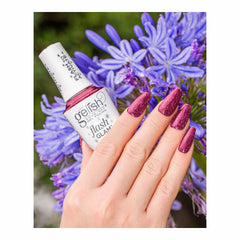 Mano con uñas vino con glitter sosteniendo el frasco blanco Gelish Flash Glam Mesmerized By You con tapa FLASH TO ACTIVATE, entre flores moradas