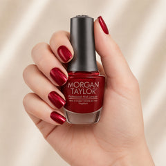 Mano sosteniendo el frasco de esmalte tradicional Morgan Taylor What's Your Poinsettia?, color Rojos acabado Shimmer, con las uñas pintadas del mismo tono